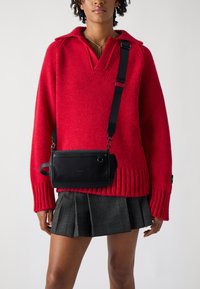 Maglione rosso a maglia con scollo a V e orlo a coste abbinato a una borsa a tracolla in pelle nera con cerniera e tracolla regolabile.