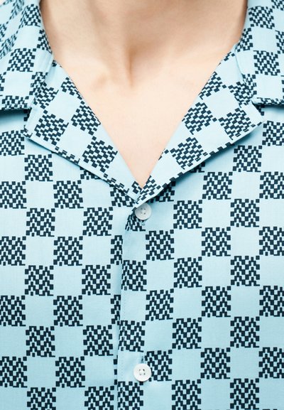 Ανοιχτό μπλε πουκάμισο με μαύρο εικονοστοιχειωτό μοτίβο "M" και ανοιχτό γιακά, που αποκαλύπτει μέρος του λαιμού και του στήθους ενός ατόμου.