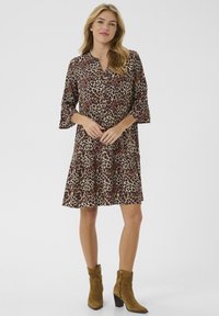 Luipaardprint jurk met een V-hals, driekwart mouwen en een gelaagde zoom. Het model draagt bruine haklaarzen. Lichte achtergrond.