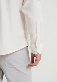 Chemise à manches longues beige clair avec une texture lisse. Poignet boutonné avec deux boutons blancs. Portée avec un pantalon gris clair, montrant un détail de poche.