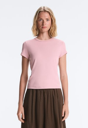 Fiatal nő vállig érő szőke hajjal, világos rózsaszín rövid ujjú pólót és sötétbarna szoknyát visel, egy egyszínű háttér előtt áll.