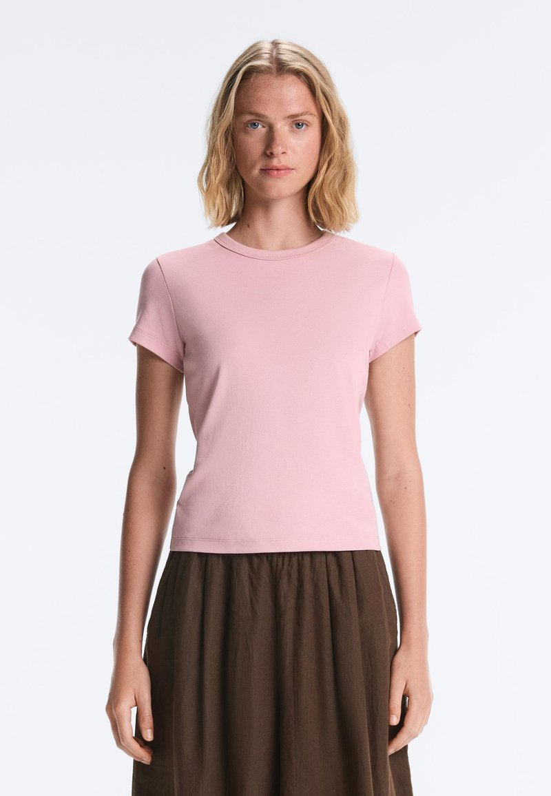Jeune femme aux cheveux blonds mi-longs portant un t-shirt rose clair à manches courtes et une jupe marron foncé, debout devant un fond uni.