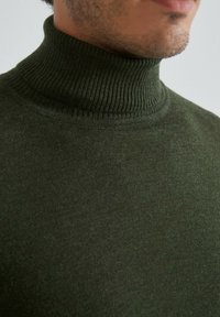 Pull à col roulé vert foncé en tissu doux, avec un col à bords côtelés et une texture lisse. Gros plan sur l'encolure et le tissu supérieur.