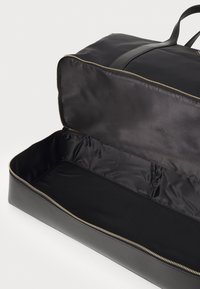 Sort duffel taske med en glat overflade, guld lynlås og rummeligt interiør foret med sort stof. Har dobbelte flade håndtag til bæring.