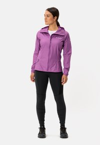 Paarse lichtgewicht jas met een ritssluiting aan de voorkant, capuchon en zijzakken; gedragen boven zwarte leggings en wandelschoenen, tegen een witte achtergrond.