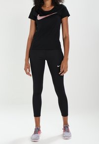 Camisa deportiva negra de Nike con un logo rosa, combinada con leggings negros y zapatos grises con acentos rosas. Tela suave, diseño ajustado.
