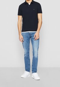 Marinblå polotröja med korta ärmar, krage och en liten logotyp; ihop med ljusblå jeans och vita sneakers. Enkelt, avslappnat design.