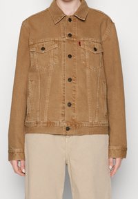 Chaqueta de mezclilla marrón con cierre de botones en la parte delantera, dos bolsillos en el pecho, bolsillos laterales y un collar puntiagudo; presenta una textura desgastada para un aspecto vintage.