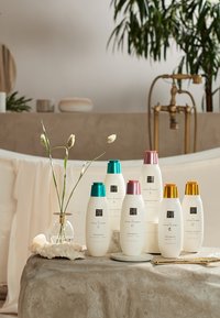 Sechs Flaschen Shampoo und Conditioner, weiß mit farbigen Kappen: grün, pink und orange. Auf einem strukturierten Steintisch mit Blumen angeordnet.