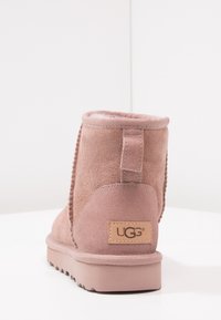 Bota UGG de ante rosa con tira en el talón y etiqueta de marca cosida en la parte trasera, mostrada desde un ángulo trasero sobre una superficie blanca.