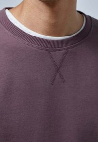 Fioletowy sweter z okrągłym dekoltem, wykonany z teksturowanej tkaniny, z prążkowanym dekoltem i szczegółem w kształcie "X" na kołnierzu, noszony na białej koszuli.