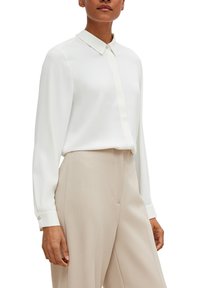 Blouse blanche à manches longues avec un col, une texture côtelée verticale et des boutons en accent sur les poignets, associée à un pantalon beige taille haute.