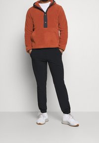 Pullover in fleece arancione con cappuccio, dotato di una patta con bottoni grigi e tasca frontale. Abbinato a pantaloni neri e scarpe sportive bianche.