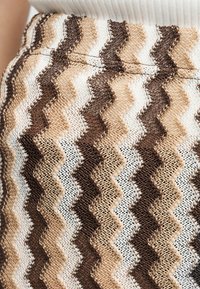 Κοντινό πλάνο υφάσματος με υφαντό ζιγκ-ζαγκ μοτίβο σε καφέ, μπεζ και λευκές αποχρώσεις σε ένδυμα που φοριέται πάνω από λευκή μπλούζα με ριμπ ύφανση.