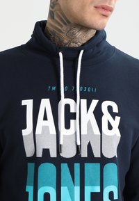 Felpa con cappuccio blu navy con colletto alto, caratterizzata da un grande design testuale bianco e turchese. Include cordoncini bianchi e un tessuto morbido e strutturato.