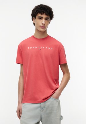 Bărbat tânăr cu păr creț și închis la culoare, purtând un tricou coral Tommy Jeans și pantaloni gri deschis, o mână în buzunar, fundal neutru.