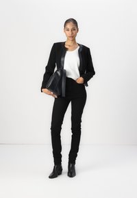 Sacou negru cu rever lucios, bluză albă, blugi skinny negri și botine. Ținând o geantă de umăr din piele neagră. Design simplu, modern.