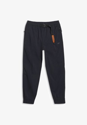 Kind met lichtbruin haar, draagt een lichtgrijze hoodie, zwarte broek met een oranje label en oranje en marineblauwe sneakers, staat met handen in de zakken.