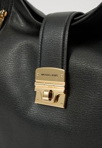 Sac à main en cuir noir avec une finition texturée, fermeture en métal doré et une large bandoulière. Le nom de la marque est embossé sur la fermeture.