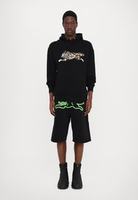 Haut à capuche noir avec un graphique de chat en imprimé léopard, associé à un short noir avec un design de contour vert. Porté avec des bottines noires à la cheville.