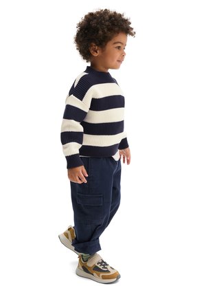 Jeune enfant aux cheveux bouclés portant un pull rayé bleu marine et blanc, un pantalon cargo bleu marine et des baskets beige et noires, debout de profil.