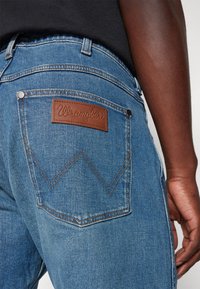 Artiškinė mėlynos Wrangler džinsų kišenė su ruda odine logotipo etiketė, dėvima asmens su tamsia oda ir juoda marškiniais.