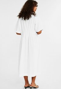 White midi dress with short puffed sleeves, textured fabric, pleated back detail, and a loose fit, paired with simple sandals.