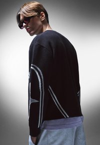 Svart stickad tröja med vita geometriska mönster, sänkt axelparti och en lager av lavendelfärgad skjorta under, i kombination med mörka solglasögon.