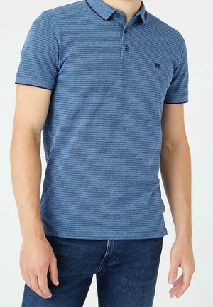 Mężczyzna w niebieskiej, krótkorękawowej, pasiastiej koszulce polo oraz ciemnoniebieskich dżinsach, pokazany od szyi do bioder.