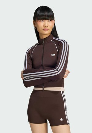 Vrouw met lang zwart haar die een bruine Adidas jas met ritssluiting en bijpassende short met witte strepen draagt, staand met gekruiste armen.