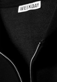 Chaqueta negra con cremallera y tejido suave, y un interior texturizado. Presenta detalles de cremallera plateada y una etiqueta blanca "WEEKDAY" en el cuello.