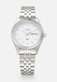 Montre en métal argenté avec un cadran blanc, une lunette striée et un bracelet à maillons argentés. Présente un affichage de la date et du jour, ainsi qu'un rétroéclairage INDIGLO.