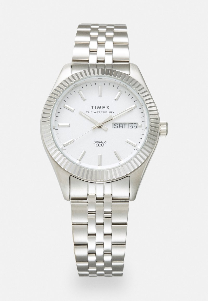 Montre en métal argenté avec un cadran blanc, une lunette striée et un bracelet à maillons argentés. Présente un affichage de la date et du jour, ainsi qu'un rétroéclairage INDIGLO.