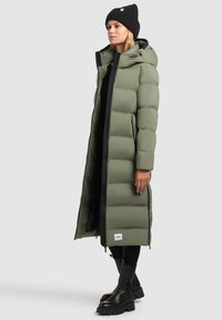 Manteau matelassé long vert olive avec capuche, présentant des sections matelassées verticales, des détails de fermeture éclair noirs et un logo en patch près de l'ourlet.