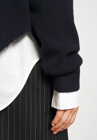 Main reposant sur le côté portant un poignet de chemise blanche, une jupe noire à fines rayures et un manche de pull en maille noire sur un fond uni.