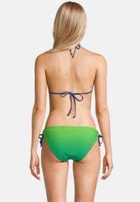 Vrouw in een blauwe halter bikinitop en groene bikini slip met zijstrikjes, gezien vanaf de achterkant tegen een eenvoudige witte achtergrond.