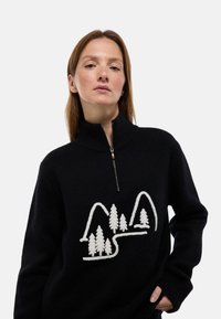 Pull noir avec un col haut, ornée d'un design brodé blanc de montagnes et d'arbres. Texture lisse et fermeture éclair.