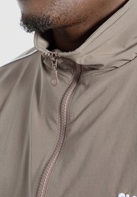 Giacca a vento leggera color taupe con colletto alto, cerniera frontale e cuciture tono su tono. Presenta una leggera lucentezza e un logo ricamato.