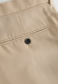 Beige Stoff mit glatter Oberfläche verfügt über eine Gesäßtasche, akzentuiert von einem schwarzen Knopf und feinen gelben Nähten.