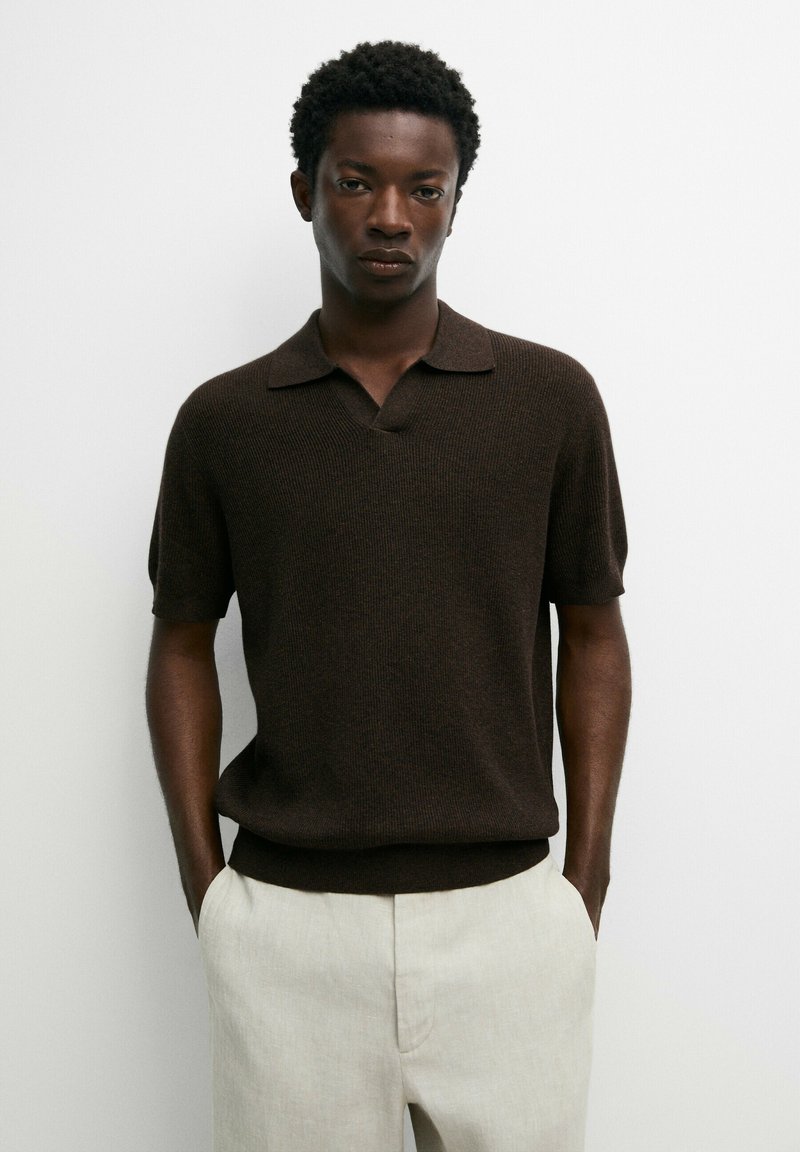 Young man with short curly hair wearing a dark brown short-sleeve knit shirt and light beige pants standing against a plain white background.