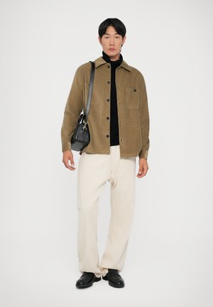 Browns korduroja jaku krekls, melna turtleneck krekls, krēmkrāsas bikses, melnas kurpes un maza melna pleca soma. Vienkārša fona.