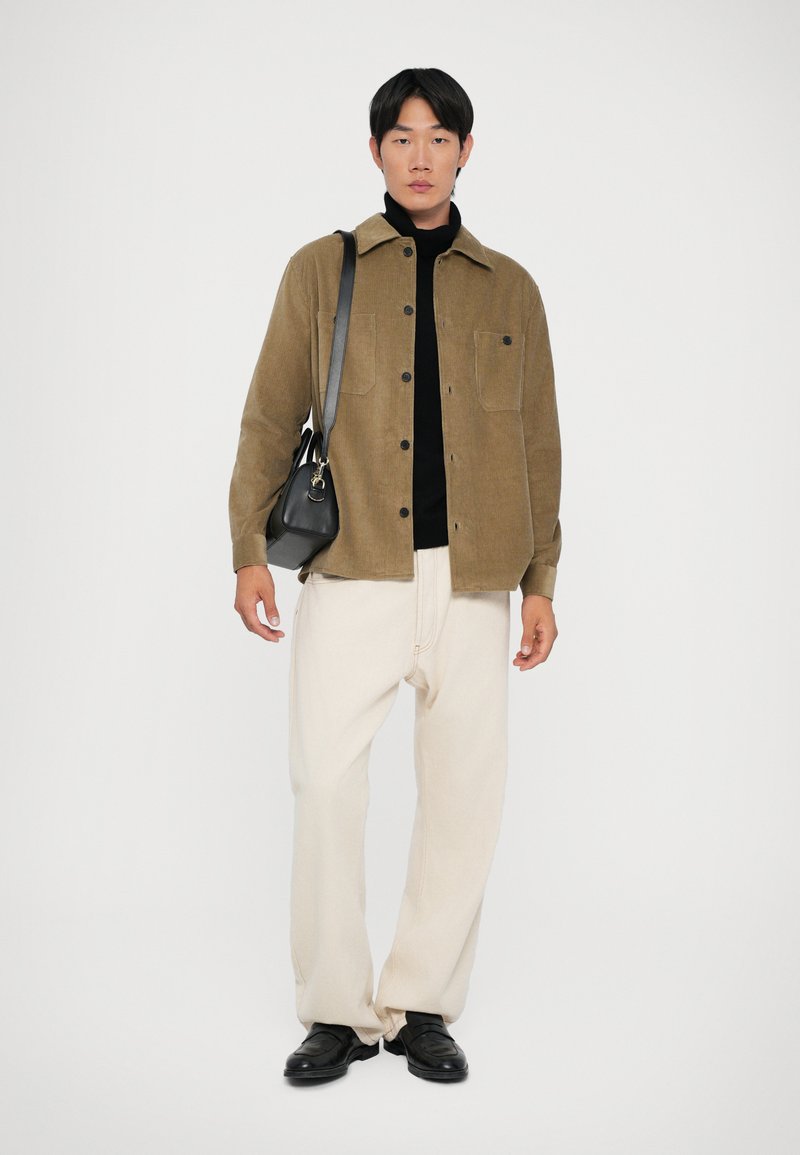 Giacca camicia in corduroy marrone, dolcevita nero, pantaloni crema, scarpe nere e una piccola borsa a tracolla nera. Sfondo semplice.