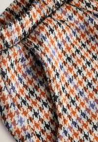Dzianina z wzorem houndstooth w kolorze pomarańczowym, czarnym i fioletowym na białym tle; miękka, strukturalna tkanina z widocznymi włóknami.