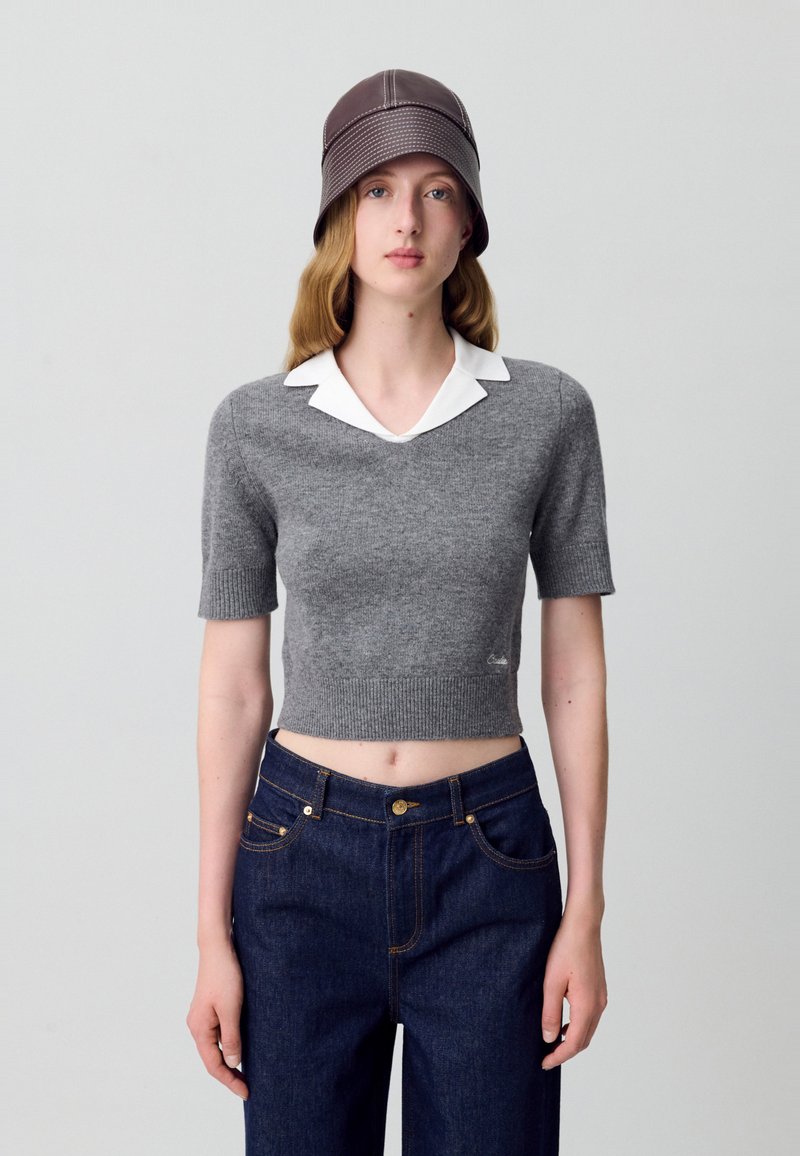 Maglione corto grigio a maniche corte con collo bianco, abbinato a jeans di denim blu scuro. Presenta una texture a costine e un discreto dettaglio del logo.