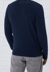 Dunkelblauer Strickpullover mit rundem Ausschnitt und gerippten Bündchen, kombiniert mit grauen strukturierten Jeans, von hinten betrachtet.