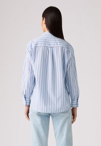Chemise à rayures bleues avec des rayures verticales rouges, à manches longues et à coupe décontractée. Elle présente un col classique et des poignets boutonnés.