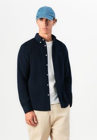 Bărbat îmbrăcat cu o șapcă albastră, cămașă bleumarin cu nasturi peste un tricou alb și pantaloni bej, stând cu o mână în buzunar, pe un fundal alb.