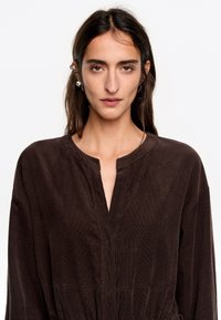 Vestido de pana marrón con mangas largas, escote en V profundo, cintura fruncida y textura sutil. Diseño sencillo sin patrones ni acentos.