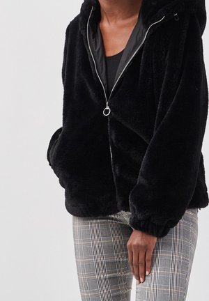 Veste noire duveteuse zippée avec tirette en anneau portée sur un top noir, associée à un pantalon gris à carreaux, main reposant sur la cuisse.