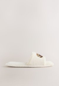 Chausson blanc en peluche à bout ouvert avec un détail brodé d’un papillon marron et doré sur la bride, vu de profil.